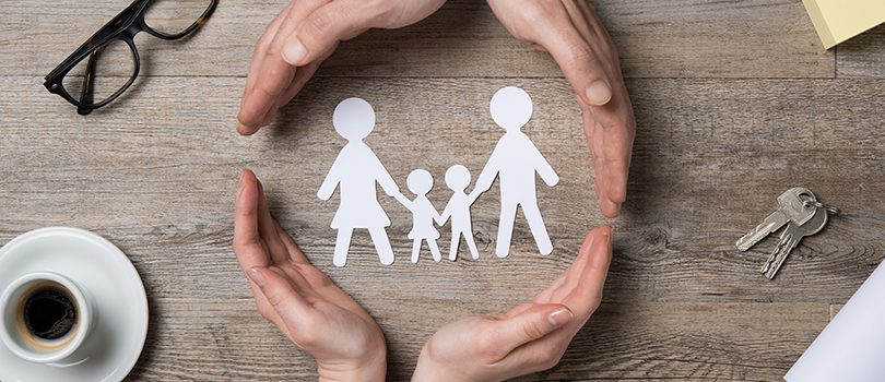 Close up of female and male hands protecting a paper chain family. Top view of two hands form a circle around white paper chain family on wooden table. Family care, insurance and helping hand concept.