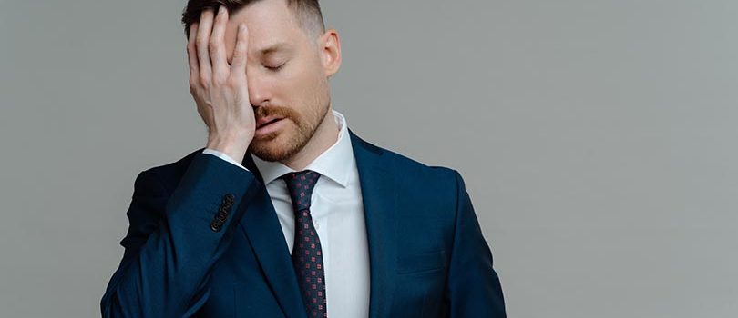 Upset dissatisfied business owner or male employee in formal wear holding hand on face and feeling frustrated and stressed of receiving bad news while standing against grey background. Failure concept